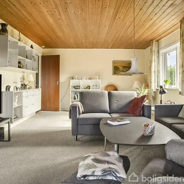 Gråt sofabord står foran en L-formet grå sofa i stue med træloft og beige vægge. Et stort fjernsyn er placeret til venstre, og vindue med gardiner til højre.