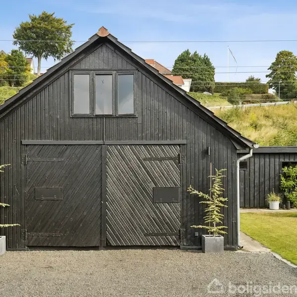 En sortmalet garagebygning med dobbeltdøre står på en grusplads. To små, slanke grantræer i potter flankerer døren. Bagved ses grønne buske og en skråning med træer.