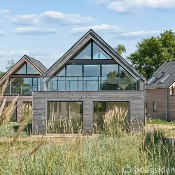 Et moderne hus med store glaspartier og trærammer står blandt højt græs. Det har en trekantet gavl og er omgivet af lignende huse og frodig natur i klart vejr.