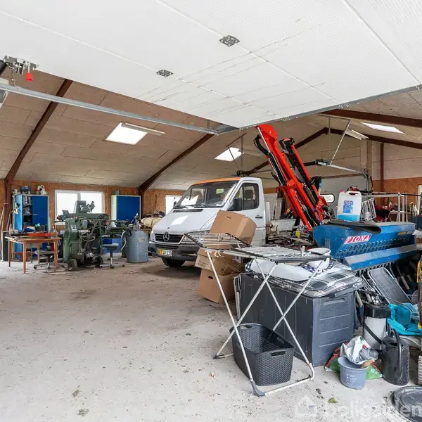 En varevogn og diverse værktøjer står i en rodet garage med skråt loft, store vinduer og arbejdsbord. Miljøet indikerer en arbejdsplads med mange genstande stablet omkring.