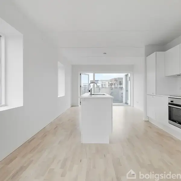 Køkken, moderne og lyst, med en central køkkenø, hvidt interiør og integrerede apparater, i en åben planløsning med store vinduer, der afslører byudsigt.