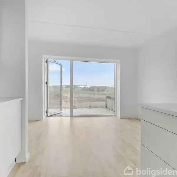 Et lyst køkken med hvide skabe og en indbygget ovn. I baggrunden er en stor glasdør, der åbner ud til en altan med udsigt over et grønt område.