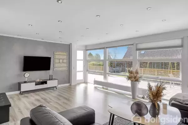 Stue med grå sofaer og et fladskærms-tv på en grå væg. Store vinduer viser udsigt til en terrasse og have. Lyst trægulv og dekorative genstande på et bord.