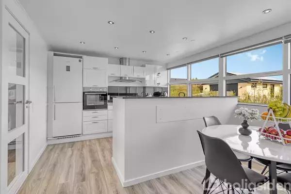 Køkken med hvide skabe og apparater, herunder ovn og køleskab, står i et lyst rum. Rundt spisebord med stole ses i forgrunden, ved siden af store vinduer med udsigt til have.