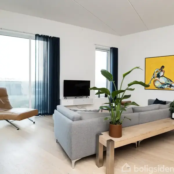 En grå sofa står i en moderne stue med en gul kunstplakat på væggen. Rummet indeholder også en brun lænestol, et sofabord med planter og store vinduer med blå gardiner.