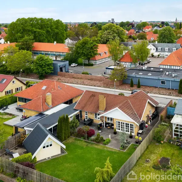 Huse med røde tage står stille i en have med grønne plæner og træer; de omgivende bygninger og et skoleområde kan ses i baggrunden.