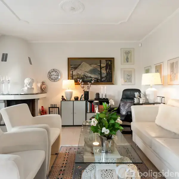 Stue med sofa og lænestol foran pejs, omgivet af kunstværker på væggen og dekorative genstande. Glasbord med blomster i mellem, tæppe og lamper skaber en hyggelig atmosfære.