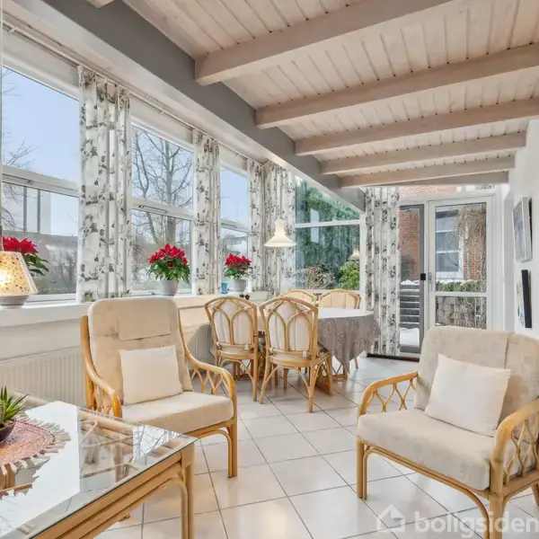 Rattanmøbler står omkring et sofabord med en plante i en lys stue med store vinduer. Baggrunden indeholder en spisebordssæt ved siden af vinduer udsmykket med røde blomster.