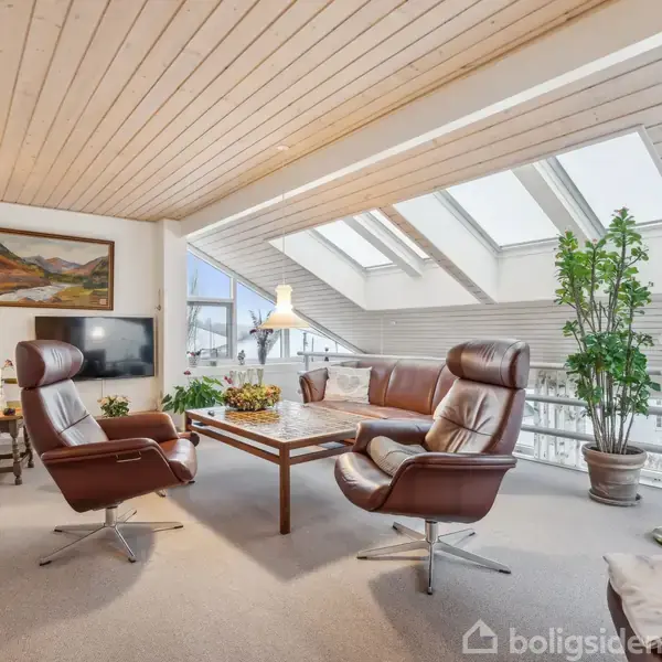 To lænestole og en sofa står omkring et sofabord i en lys stue. Skråvinduer i loftet slipper naturligt lys ind. Indretningen er klassisk med planter og kunst.