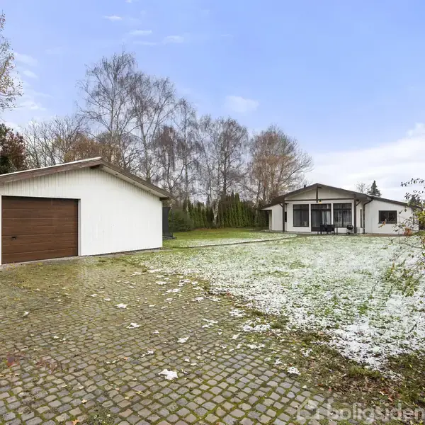 En hvid bygning med garage er til venstre, mærket med "29". En villa med terrasse ligger til højre. Beplantning og træer omringer området; jorden er delvist dækket af sne.