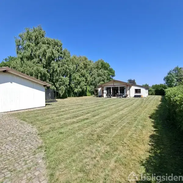 Hus med overdækket terrasse står for enden af en velplejet græsplæne, omgivet af grønne hække og træer. Til venstre ses en hvid garage med stenbelagt indkørsel.