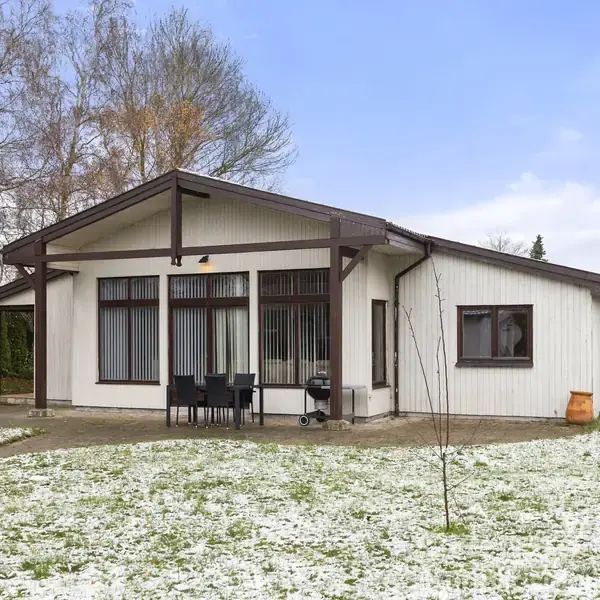 Et hvidt træhus med store vinduer står i en have med let snefald. Foran huset er et bord med sorte stole. Træer omgiver ejendommen i baggrunden.