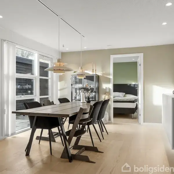 Spisebord med seks stole står i et lyst rum med trægulv. Store vinduer til venstre, og en åben dør fører til et soveværelse. Vægge har kunst og et ur.