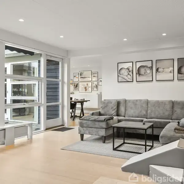 Grå hjørnesofa står i en lys stue med lyse trægulve, ved siden af et sofabord. Store vinduer giver udsigt til en terrasse. Væggen prydes af fotografier.