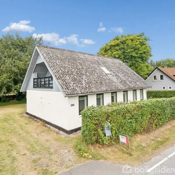 Et hvidt hus med skråtag står ved en vej omgivet af en hæk. Til venstre ses en græsplæne og træer. I baggrunden står endnu et hus.