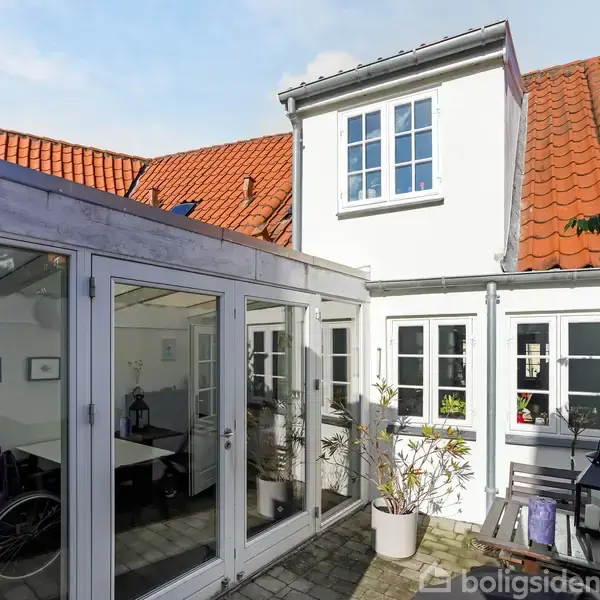 Hvidt hus med rødt tegltag, vinduer og glasdøre åbner op til en lille indre gårdhave. Sollys skinner ind, og blomster i potter pryder området.