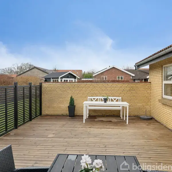 Træterrasse med hvid bænk og bord op ad gult murstenshus, omgivet af sort hegn. Blomsterdekoration på mørkt bord i forgrunden. Enkel familiehusbaggrund, blå himmel.