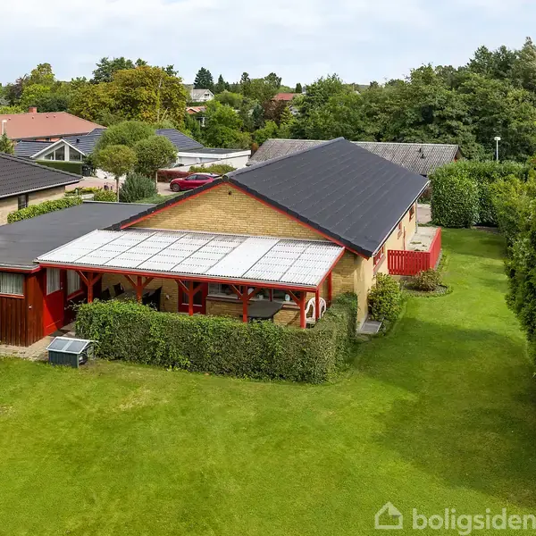 En gul murstensvilla med sort tag har en overdækket terrasse og er omgivet af en grøn have med hække. Den ligger i et parcelhuskvarter med flere lignende huse.