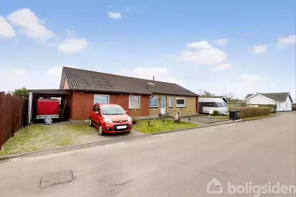 En rød bil holder parkeret foran et murstenshus med fladt tag. Huset har en carport og står i et villakvarter med en campingvogn til højre.