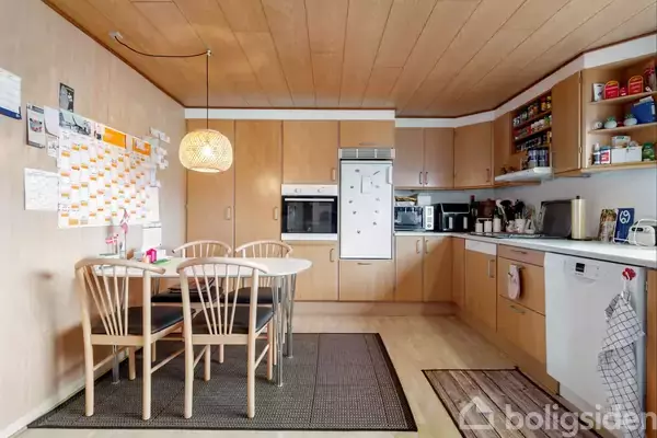 Et køkken med træindretning har et spisebord med fire stole under en hængende lampe. Køkkenet indeholder en ovn, opvaskemaskine, og hylder med opbevaring. En stor vægkalender ses på væggen.