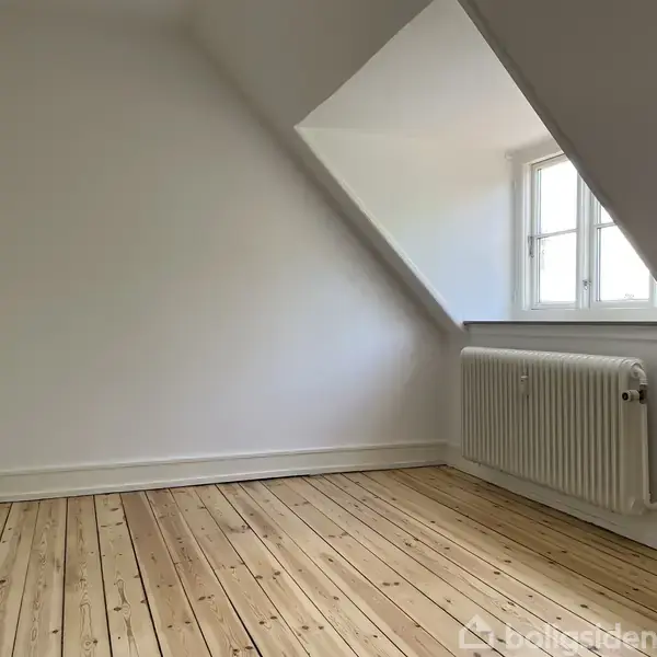 Et værelse med hvidmalede vægge og et trægulv. Der er en radiator under et skråvindue, som lader sollys komme ind i rummet.