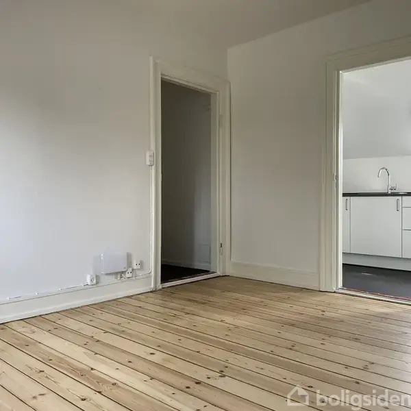 Et tomt rum med lakeret trægulv fører til et tilstødende køkken med hvide skabe og sort bordplade. Væggene er hvide, og der er en døråbning mellem rummene.