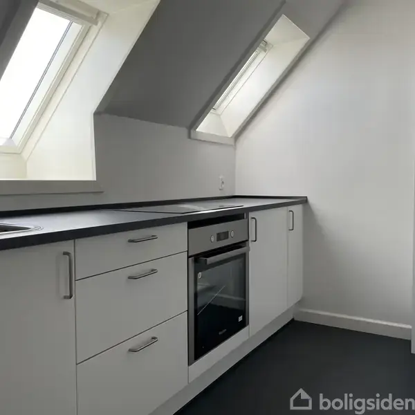 Et moderne køkken med hvide skabe og sorte bordplader. Ovnen er indbygget. To ovenlysvinduer giver naturligt lys. Rummet er minimalisktisk og lyst.
