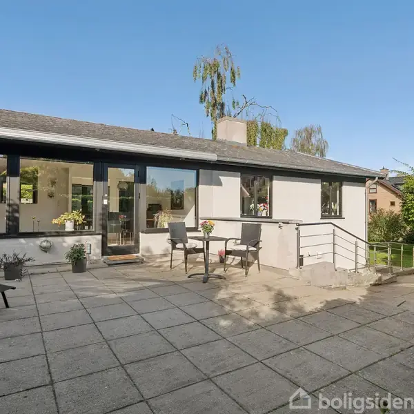 Hus med store vinduer, hvor der står havemøbler på en flisebelagt terrasse. Omkranset af grønne buske og træer, under en klar blå himmel.
