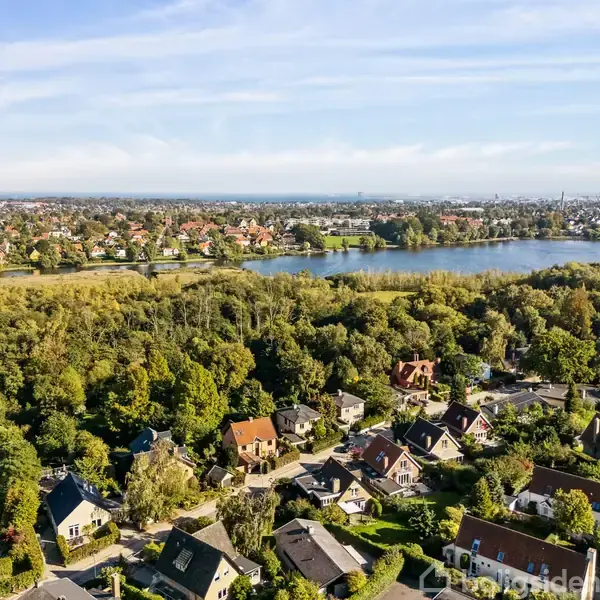 Huse ligger spredt blandt grønne træer. Over dem findes en sø, der strækker sig gennem landskabet. I baggrunden ses en by med flere bygninger.