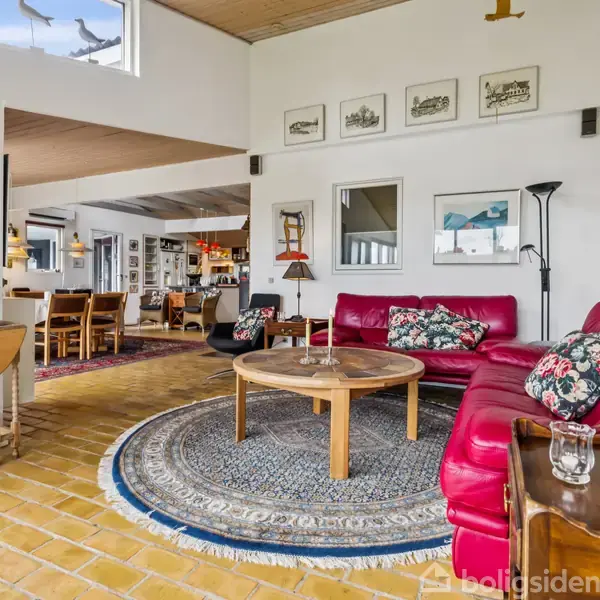 Rød sofa står i stue, omgivet af et rundt tæppe og træmøbler, med pudebetrukne puder. Væggene har flere indrammede billeder, og rummet fører ind til en spisestue med spisebord.