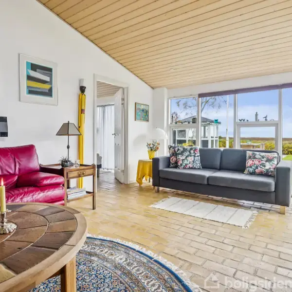 Stue med rød og grå sofa dekoreret med farverige puder, placeret på klinkegulv. Store vinduer mod en haveudsigt. Væggene er udsmykket med moderne kunst. Rundt sofabord med lys.