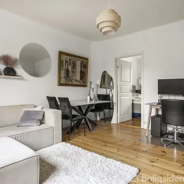 En lys stue har sofa til venstre, et rundt spisebord med stole i midten og et skrivebord med computere til højre. Der er trægulv og hvidmalede vægge.