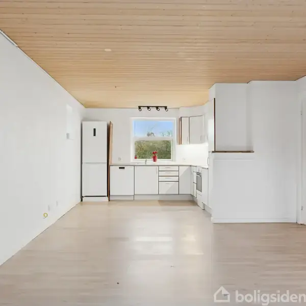 Køkkenet har hvide skabe og køkkenapparater, med en stor vindue i baggrunden. Rummet er lyst med trægulve og træloft, omgivet af hvide vægge.