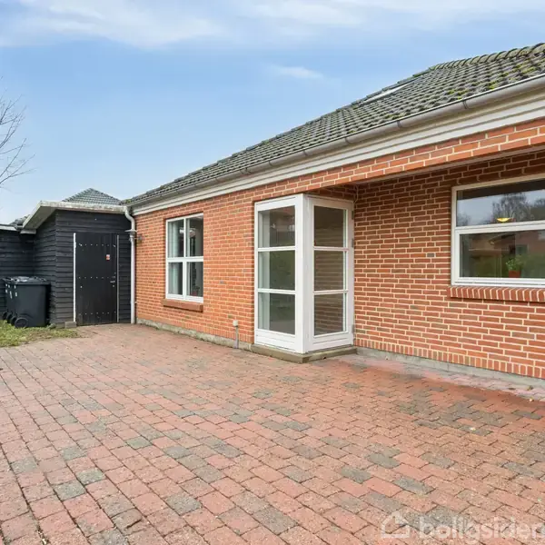 Rødt murstenshus med store vinduer står stille. Foran er en bred flisebelagt terrasse. Til venstre ses en sort skurbygning. Himlen er skyfri, og der er træer i baggrunden.