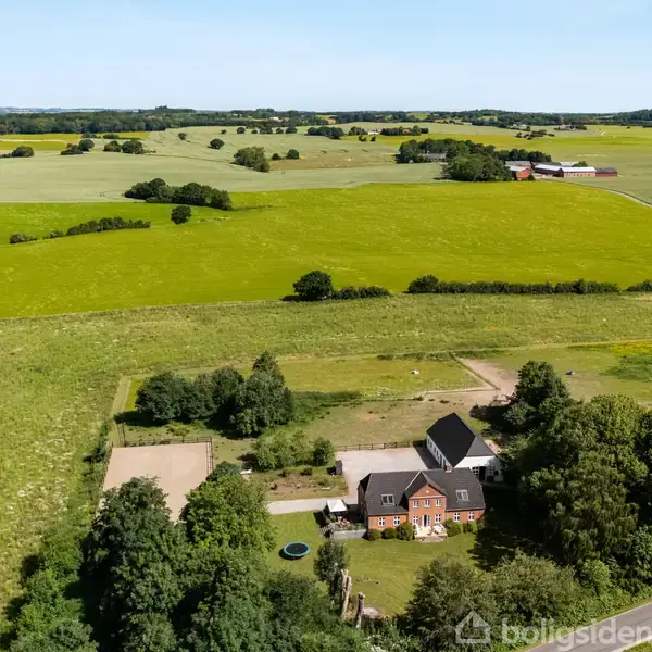 En stor, rød murstensbygning omgivet af frodige marker og træer ligger i et åbent landdistrikt. Landbrugsarealer og spredte træer strækker sig i baggrunden under en klar, blå himmel.
