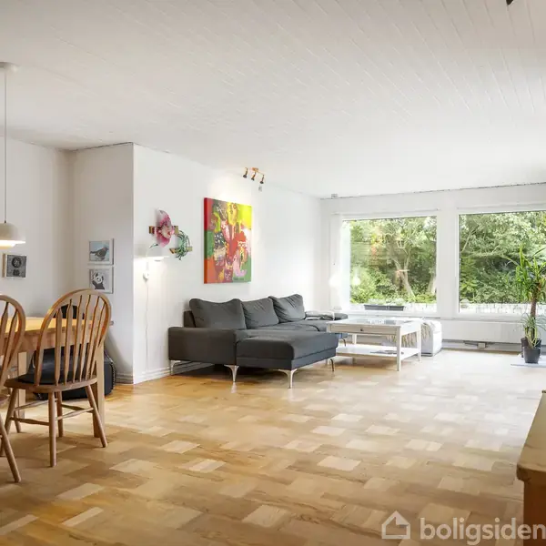 Stue med grå sofa ved siden af et stort vindue, der giver udsigt til en grøn have. Et spisebord med stole står til venstre, og væggene har kunstværker.