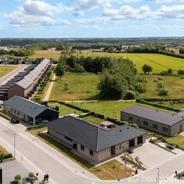 Boligområde med moderne huse placeret langs lave gader. Husene har mørke tage, og området er omgivet af grønne marker og træer under en skyet himmel.