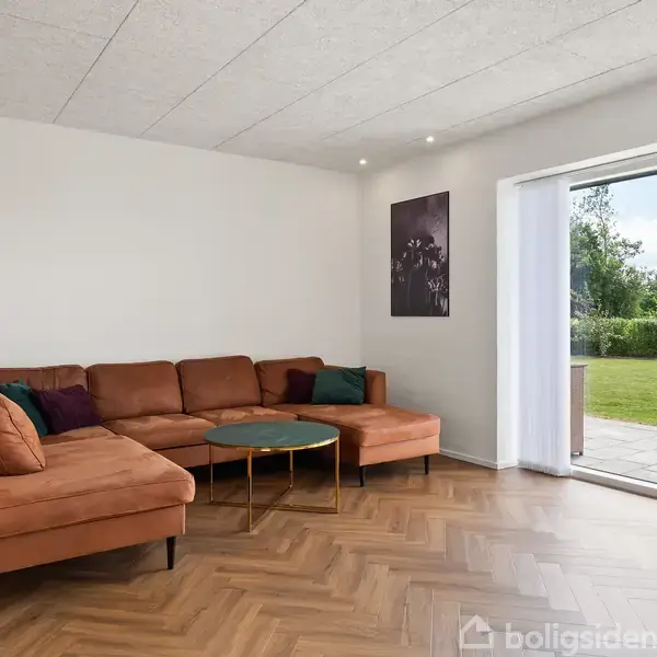 Brun hjørnesofa med puder står i stuehjørne. Rundt sofabord foran. Åben glasdør til terrasse med udsigt til have. Trægulv og minimalistisk indretning med hylder på væggen.