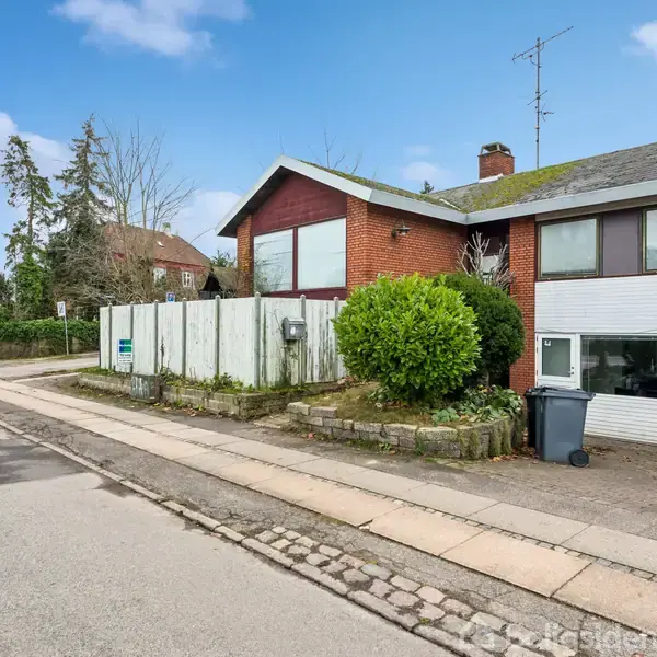 Et rødt murstenshus med store vinduer står stille langs en stille villavej. Foran huset er der en skraldespand og en stor grøn busk. Fortovet løber parallelt med vejen.