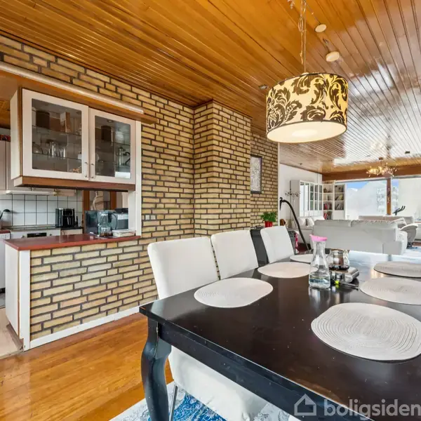 En spisestue med et sort træbord og seks lyse polstrede stole. En lampe med mønstret skærm hænger over bordet. Til venstre ses murstensvægge og køkkenskabe.