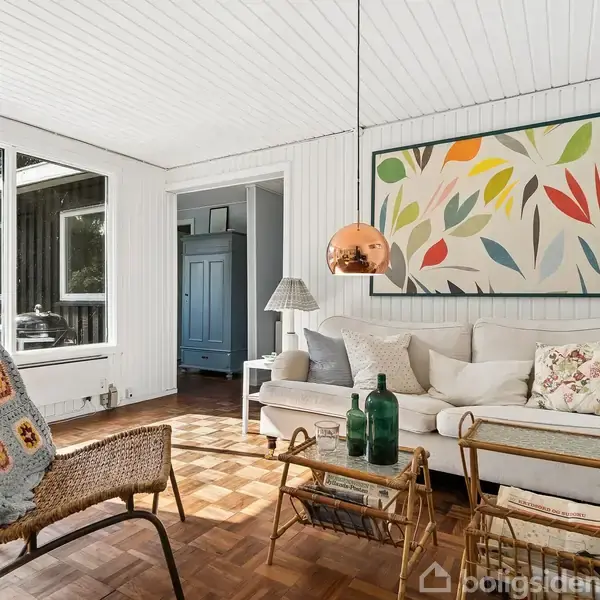 En stue med en hvid sofa fyldt med puder står foran et farverigt blomstermaleri. En gyngestol med en strikket plaid er nær ved. Rummet har lyse trægulve og store vinduer, der lukker naturligt lys ind.