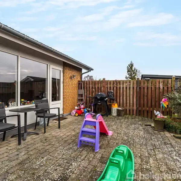 Terrasse med to sorte stole ved et stort vindue; farverige børnelegetøj spredt på fliserne; hævede træhegn og buske omkring; gråblå himmel ovenfor.