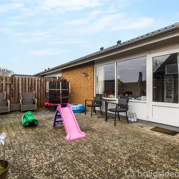 En udendørs terrasse med en lyserød rutsjebane og et legehus i en flisebelagt gård. To lænestole står ved et plankeværk, mens et bord og stole er placeret ved husets indgang.