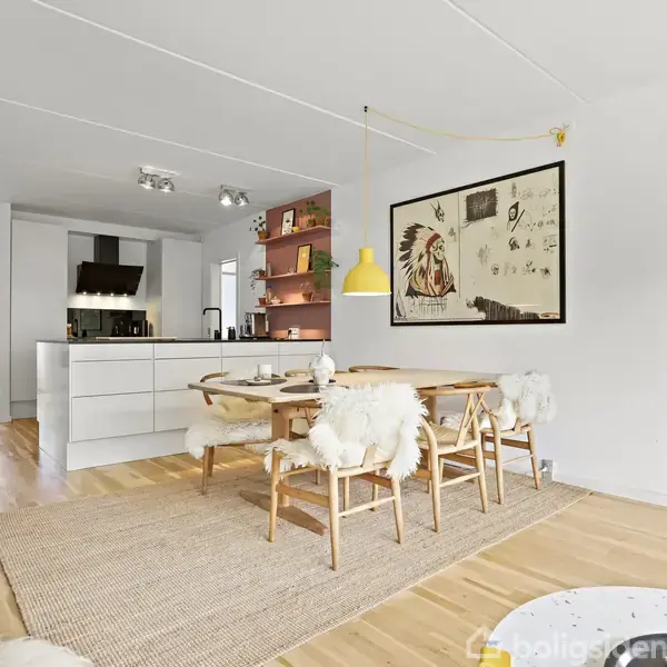 Moderne spisestue med træbord og stole med lyse hynder. Væglampe hænger over bordet. I baggrunden ses et åbent køkken med hvide skabe. Kunstværk på væggen.