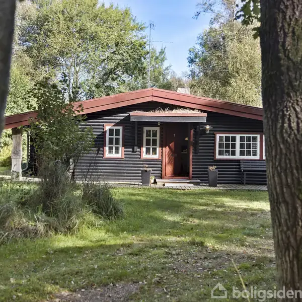 Sort træhus står stille i en frodig have omgivet af høje grønne træer. Husets røde tag og dør skaber kontrast mod vegetationen.