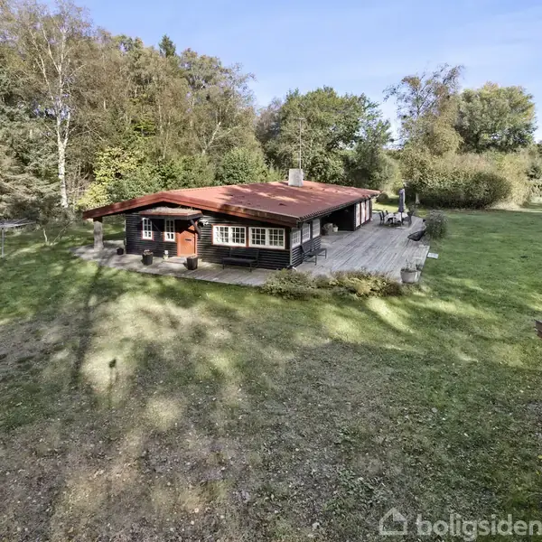 Et træhus med rødt tag står midt på en grøn plæne omgivet af skov. En stor terrasse omkranser huset, og solen skinner gennem træerne.