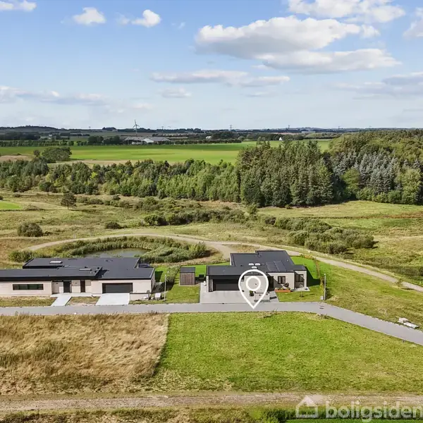 Huset står stille på en parcel med en markeret placering, omgivet af åbne marker og skovområder under en klar blå himmel med spredte skyer.