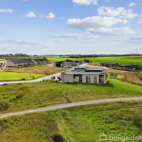 Et moderne hus med fladt tag står i en grøn, åben landlig omgivelse. En smal grusvej fører til bygningen, omgivet af marker og flere lignende bygninger i baggrunden.