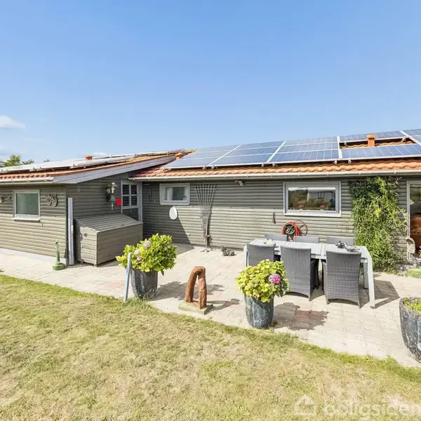Hus med solpaneler på taget står stille på en flisebelagt terrasse med planter i krukker. Græsplæne omgiver huset under en blå himmel.
