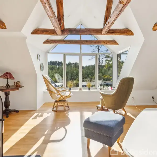 Brændeovn tændt ved siden af moderne stue med store vinduer, der lukker sollys ind. Træmøbler dekorere rummet, og udenfor ses en frodig skovudsigtsbaggrund.